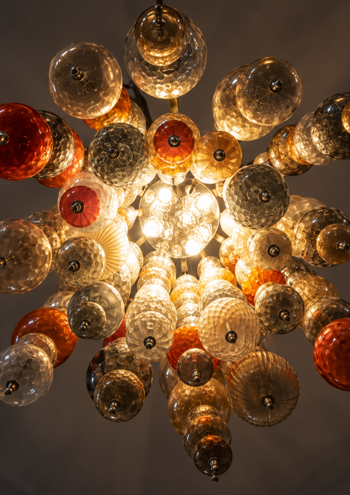 A multi-light chandelier featuring numerous textured glass globes and teardrop shapes in amber, orange, red, and clear glass, arranged in a radial starburst pattern against a dark gray background.