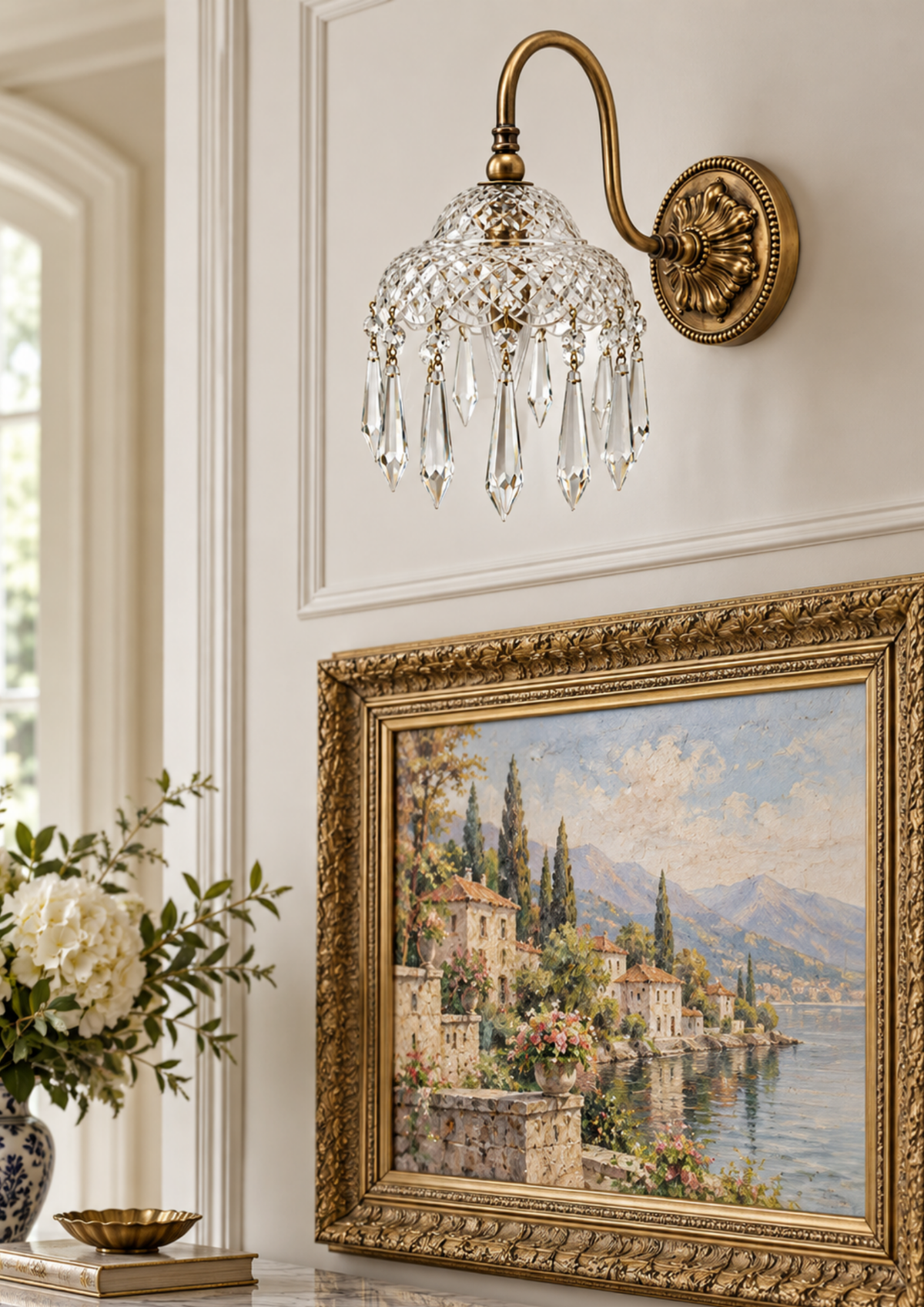 An ornate brass wall sconce with hanging crystal prisms mounted on a cream paneled wall above a large framed landscape painting.